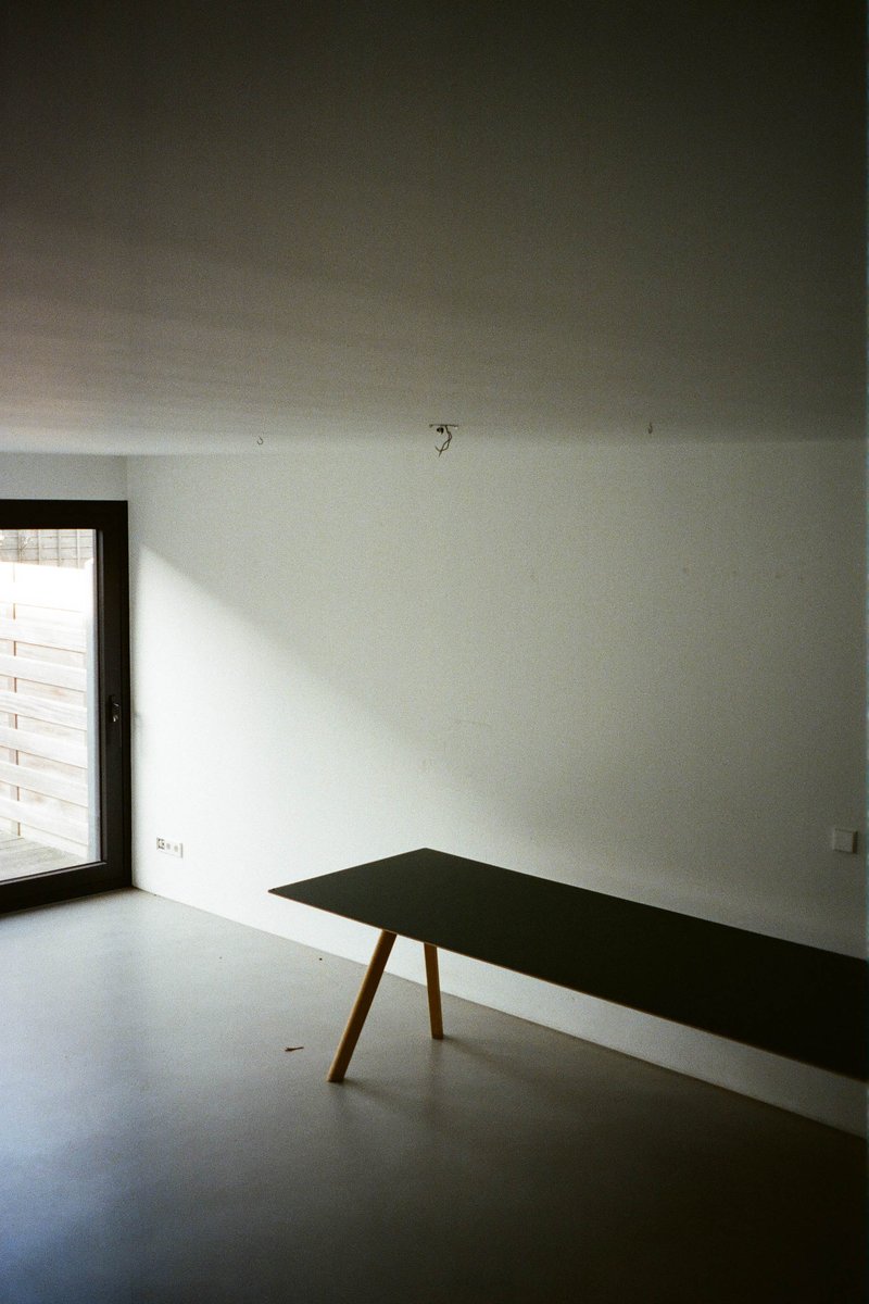 The image shows an interior space with minimal decor. Dominating the scene is a modern, minimalistic living room area characterized by clean lines, muted tones, and sparse furnishings. In one corner, there's a small, dark table with a few items on it, including what appears to be a book or a box, along with some other objects that are not clearly identifiable due to the angle and focus of the photograph. The floor is a light color, providing a neutral backdrop to the scene. The room has walls painted in shades of white and gray, contributing to the overall calm and uncluttered atmosphere.