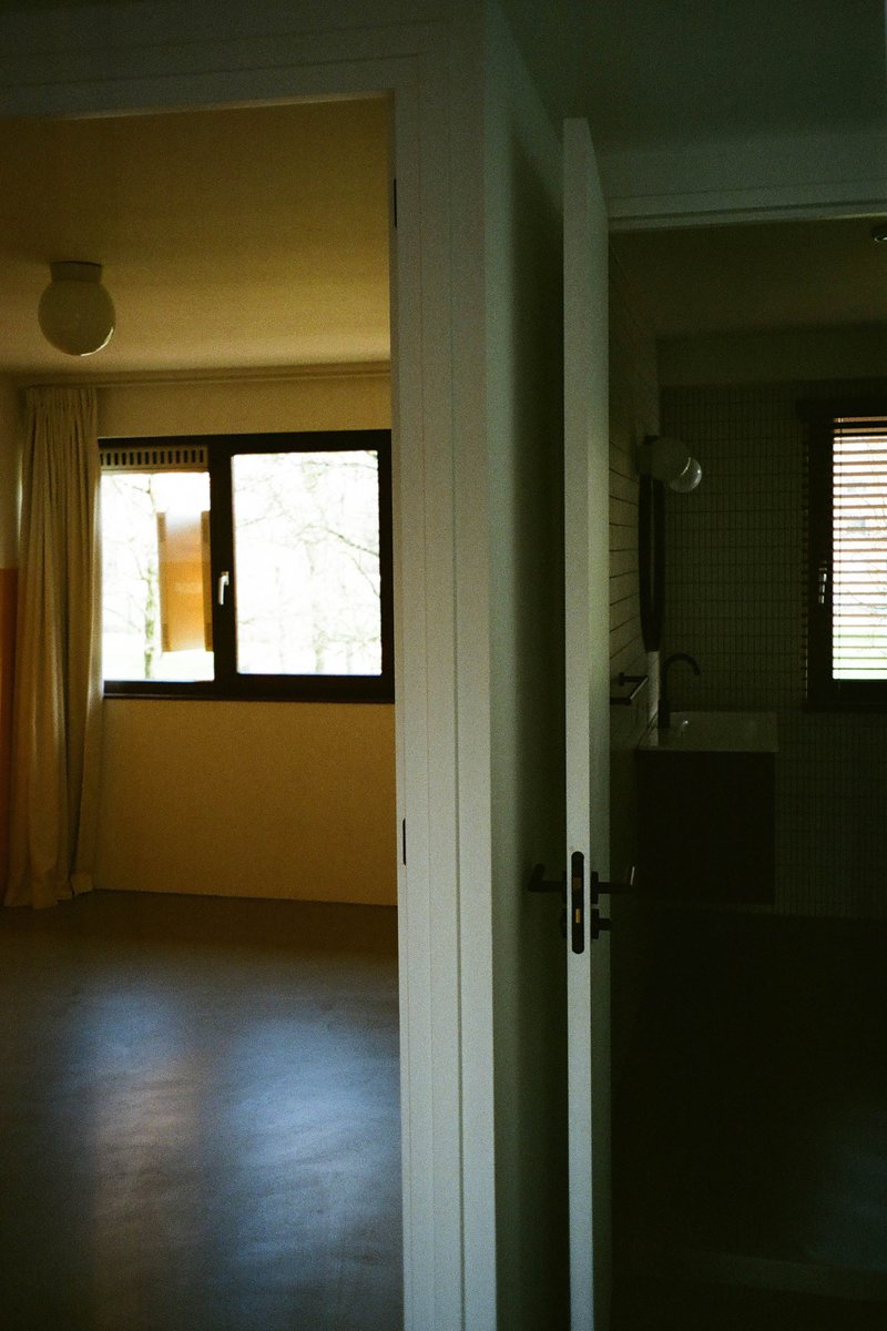 The image shows an interior space that appears to be a room with white walls and dark flooring. There are two long windows on the right side of the photo, each allowing natural light into the room. On the left, there is a large doorway leading to another room or hallway. The room has a simple, functional design, with no visible decorations or furniture that could provide additional context about its use.