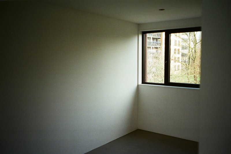 The image depicts an interior space with a white, untextured wall. In the corner of the room, there is a window with a visible shadow on it from a nearby light source. The floor appears to be carpeted and colored darker than the wall. There are no other significant objects or details that stand out in this setting.