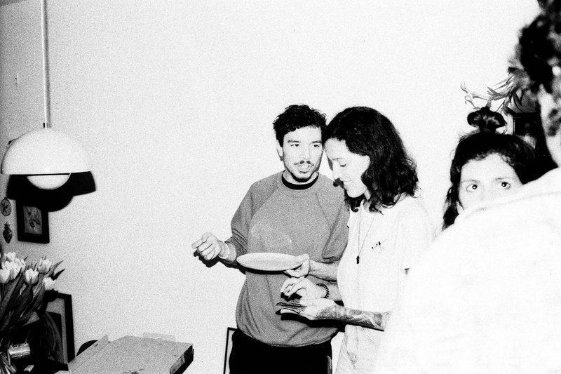 In the black and white photo, two people are standing side by side in a room with a plain wall. They appear to be engaged in a casual conversation or interaction as they hold what could be cups or small bowls in their hands. There's also a table visible in the background, but it's not clear if anything else is on it.