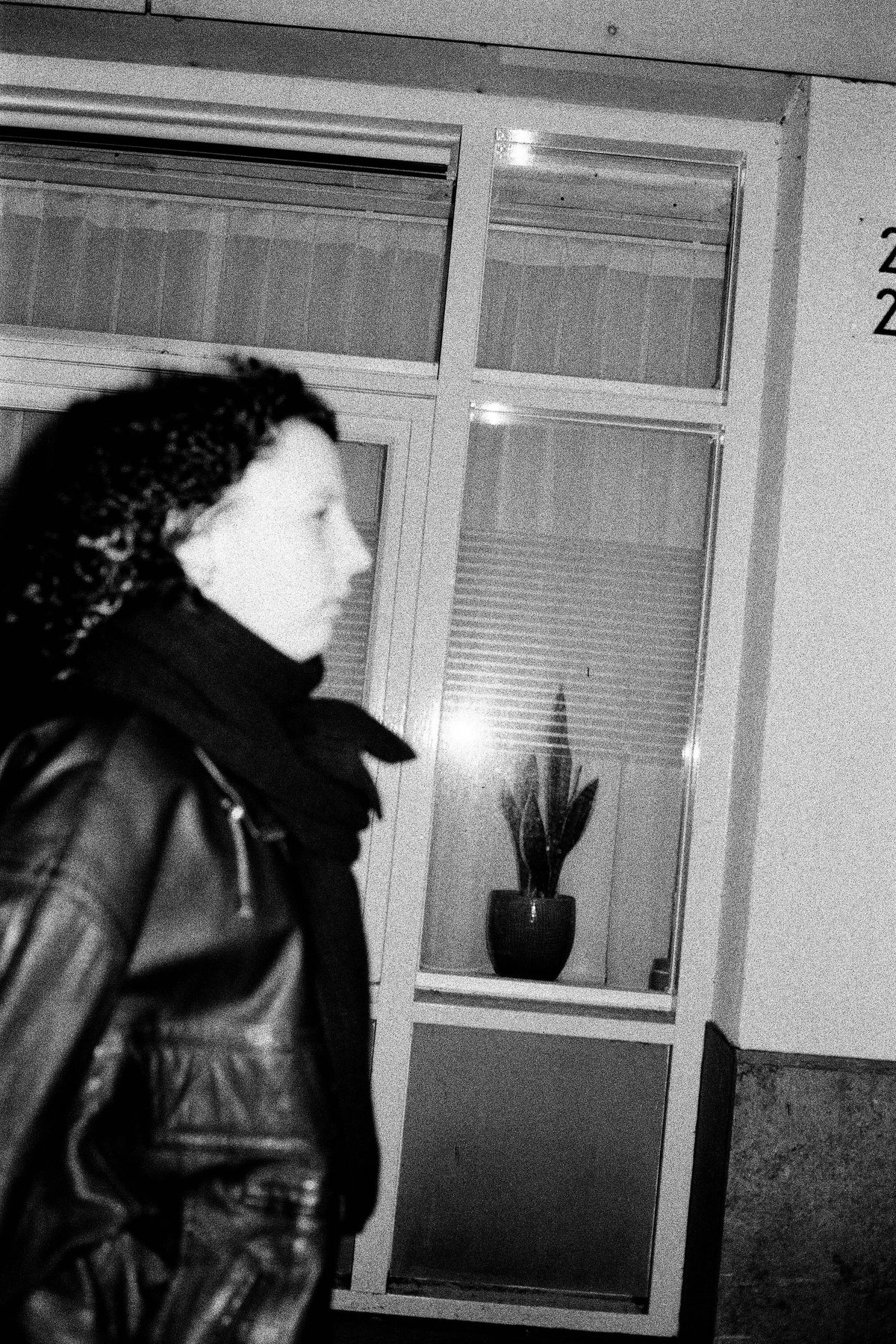  The image features an individual walking past a glass door with a reflective surface, visible behind them. They appear to be wearing dark clothing and are looking downward as they move. The overall scene is blurred, which may limit the identification of specific details or objects. 