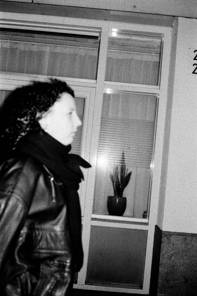 The image features an individual walking past a glass door with a reflective surface, visible behind them. They appear to be wearing dark clothing and are looking downward as they move. The overall scene is blurred, which may limit the identification of specific details or objects.