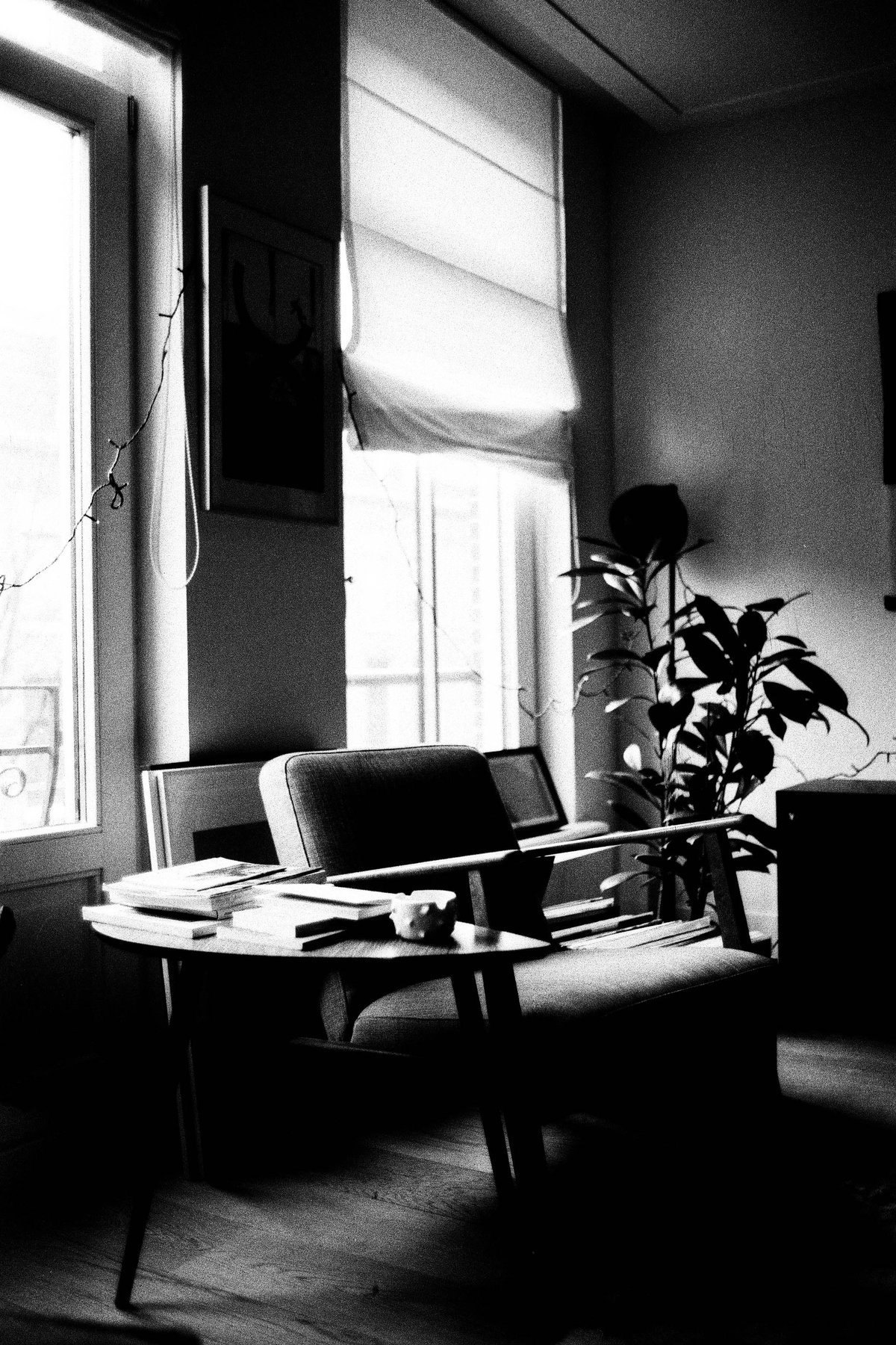  The image shows an interior space that appears to be a living room or den. In the foreground, there's a couch with a person sitting on it; behind them is a chair facing away from the viewer. There are two windows visible, one covered by blinds and the other not. A side table stands between the couch and the chair, holding a couple of items. The room has simple, muted decorations that give it a calm ambiance. 