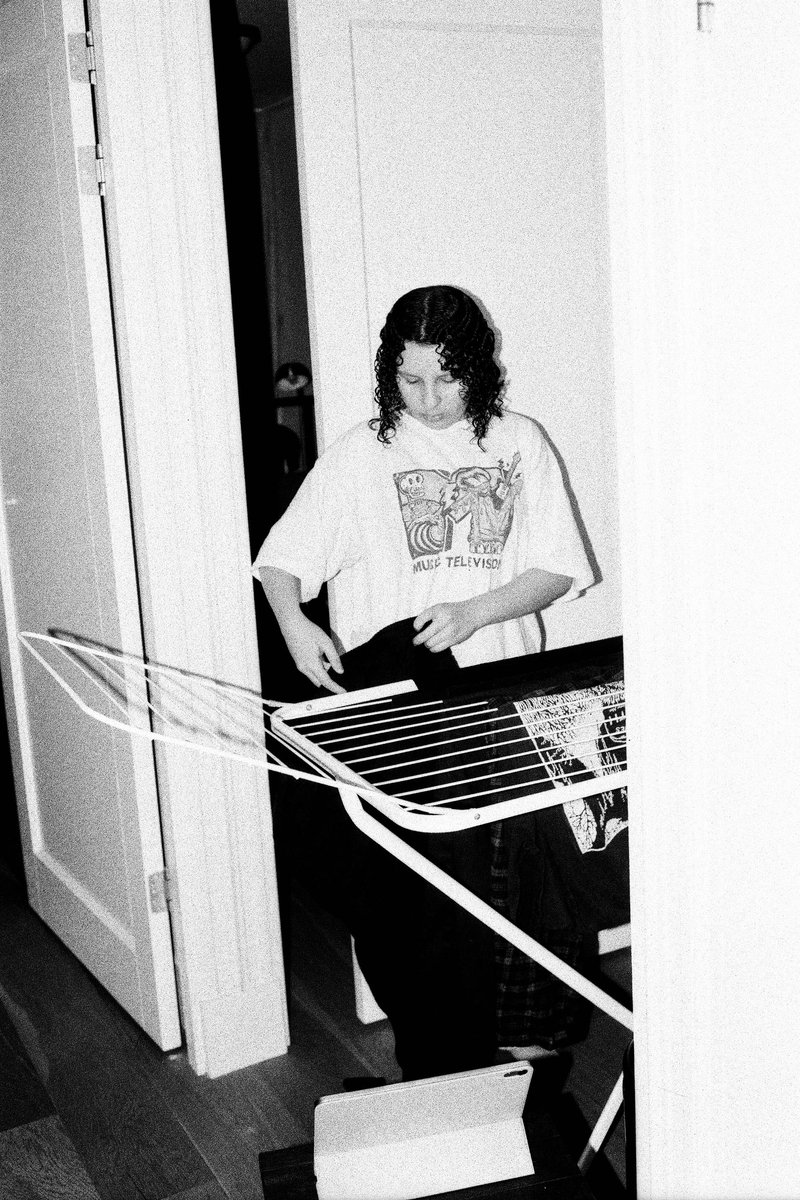 The image is a black and white photograph of a person seated near a guitar in what appears to be a domestic setting, possibly a kitchen or living room. The individual is partially visible, with only the upper body and head showing, and seems to be playing an instrument, though no details are visible due to the resolution. There are also several items around, including a table with various objects on it and a window with light coming in.