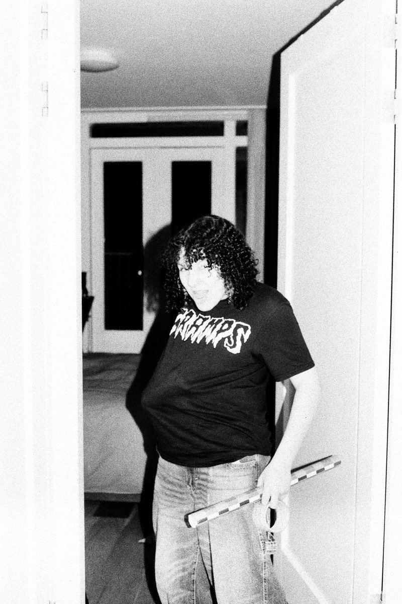 The image is a black and white photo featuring a person standing in an indoor setting with what appears to be a window behind them. The individual is wearing a t-shirt with visible text and is positioned next to an open door or possibly a hallway that leads into another room.