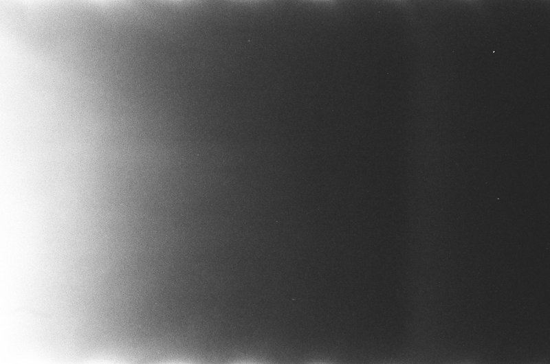 The image shows a monochromatic photograph with a blurred, high contrast quality. It appears to be a close-up of textured fabric or paper, possibly with a pattern or design that is not distinct due to the focus on the central black area. This dark void creates an optical illusion known as the "Mach bands," where the human visual system perceives an image as being lighter at the edges and darker in the center, causing the peripheral areas to seem brighter relative to the center.