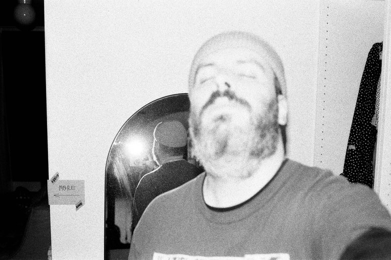 The image is a grayscale photograph that appears to show a man sitting indoors with a bald head and a beard. He's wearing a dark top, and there's a blurry background that looks like a domestic setting. There are no visible texts or distinctive objects in the photo.