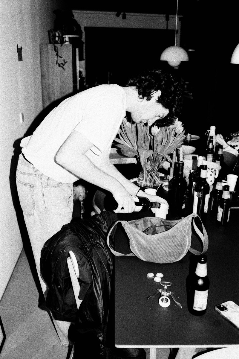 In the center of the room, a man is sitting at a table, preparing food or drink. He seems to be holding something in his hand, possibly an ingredient or a utensil for cooking or serving. The room has various objects like bottles and bowls on the table and a cup nearby. The lighting suggests it could be evening or nighttime.