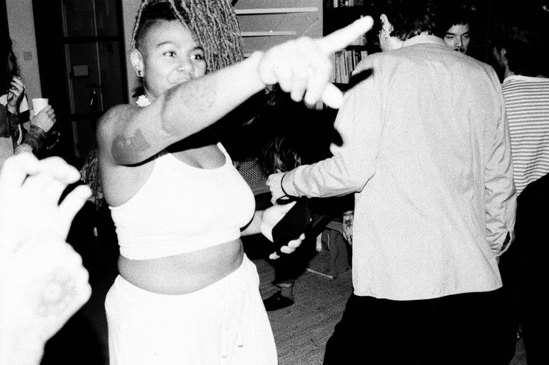 The image is a vintage black and white photograph capturing an intimate moment between two people at what appears to be a party or gathering. In the foreground, there's a woman with her arm extended, pointing towards someone off-frame. She's wearing a sleeveless top and has a focused expression on her face. Behind her, there's another person facing away from the camera, also seemingly engaged in the conversation. They are dressed in casual attire, and their position relative to the woman suggests they might be dancing or moving to the music. The background is blurred but hints at a festive atmosphere with party lights and possibly other guests out of focus.