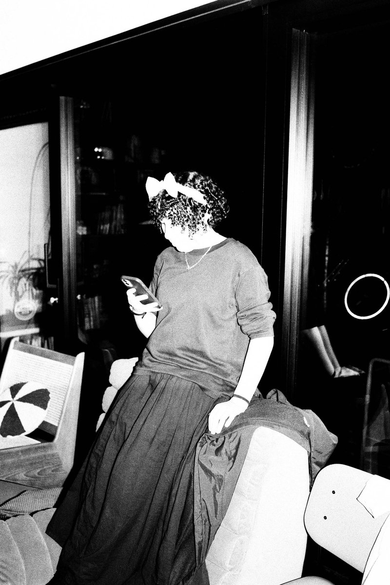 The image is a black and white photograph of a woman seated at a window. She appears to be engrossed in her cell phone, which she holds in her hands. The room has a cozy feel with a couch visible behind her. The style of the photograph suggests it might have been taken during a casual moment captured by someone else.