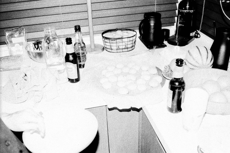 The image shows a kitchen scene with various items scattered across the countertops. There are multiple bottles of different shapes and sizes, some cups, and a few wine glasses. The overall impression is of a busy kitchen space with various items used or ready for use.