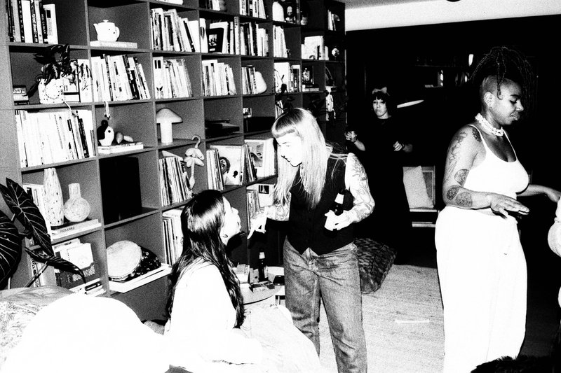 The image appears to be an older photograph, possibly taken indoors, showing multiple people engaged in what seems to be a social event or gathering. There are at least five individuals present; they seem to be in the midst of conversation or activity. One person is holding a bottle, suggesting that drinks may be involved. In the background, there's a bookcase and some decorative items on shelves, indicating a homey environment. The overall atmosphere suggests a casual, possibly celebratory gathering among friends or acquaintances.