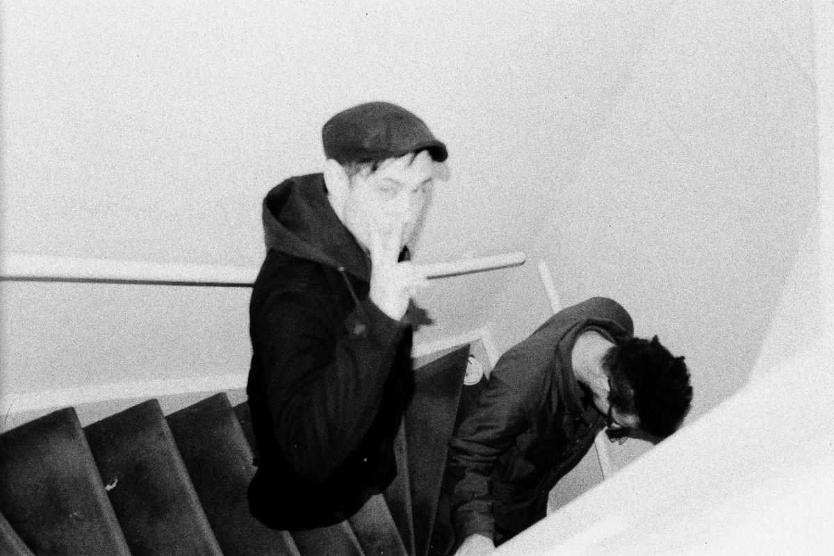 The image appears to be a black and white photograph of two individuals on a staircase. The person on the left is looking towards the camera with thei