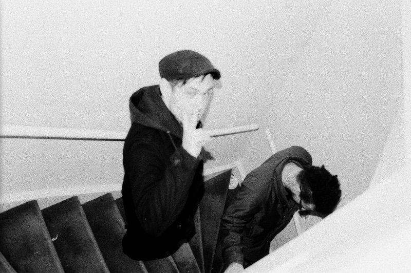 The image appears to be a black and white photograph of two individuals on a staircase. The person on the left is looking towards the camera with thei