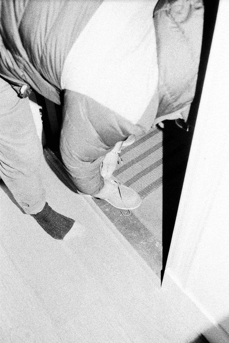 The image shows a person standing inside a doorway, looking down at the ground. They are wearing a jacket with the hood up and dark pants. The interio