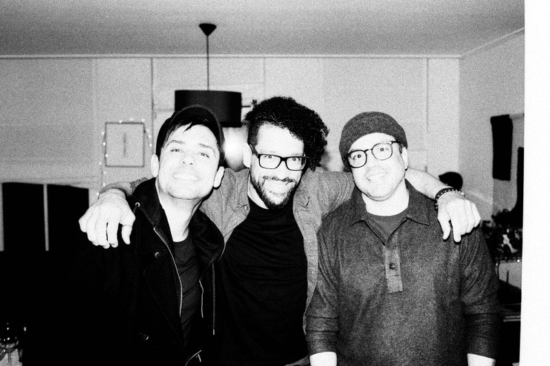 The image shows three men standing together and smiling. The central figure is more prominent due to his larger size in the frame, and he's wearing gl