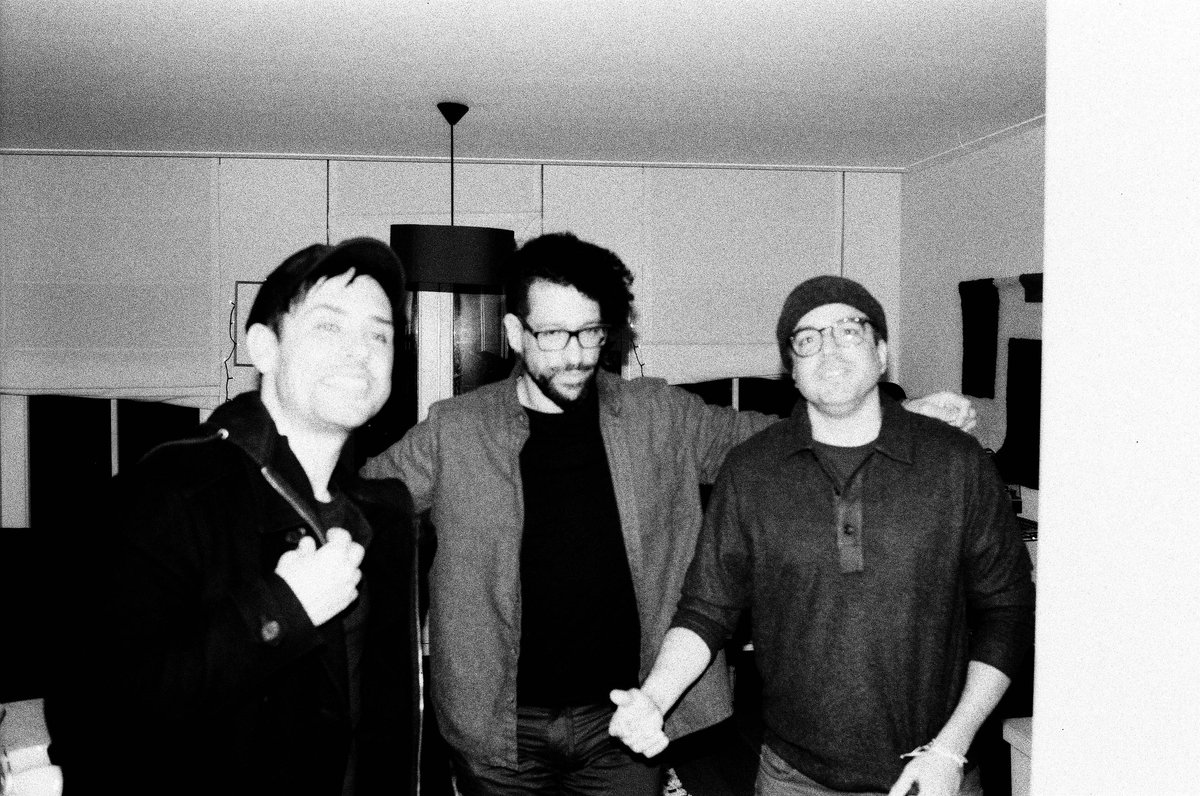 Black and white photo of three men standing together, smiling. They're in a room with a blurred background, giving focus to the subjects. The man on t