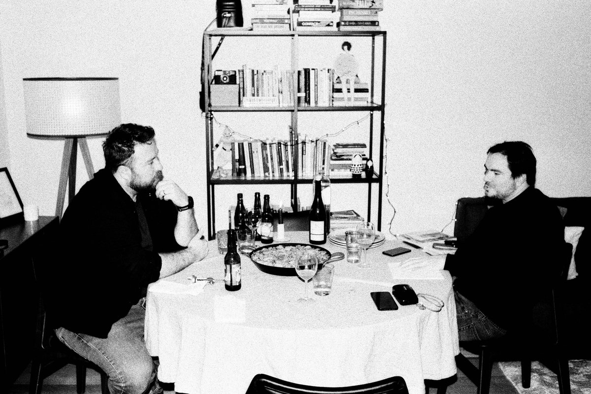 A sepia-toned image of two men at a dining table. They appear to be engaged in conversation, with one man looking up at the other. The room has a cozy