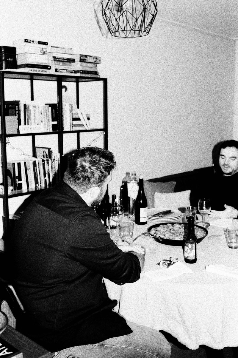 This is a black and white photo featuring two men sitting at a dining table. One man appears to be holding something up, possibly looking at it closel