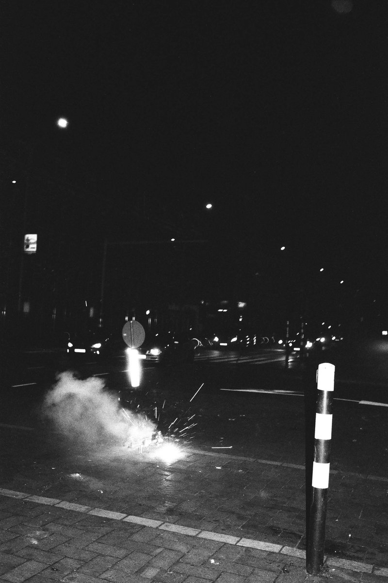 The photo captures a night scene on a city street. In the center of the image, fireworks are being set off, casting a bright light and creating a sens