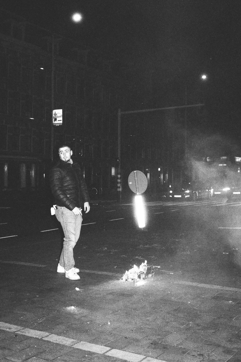 This is a black and white photograph capturing a person standing in the middle of a street, surrounded by smoke from burning trash. The person appears