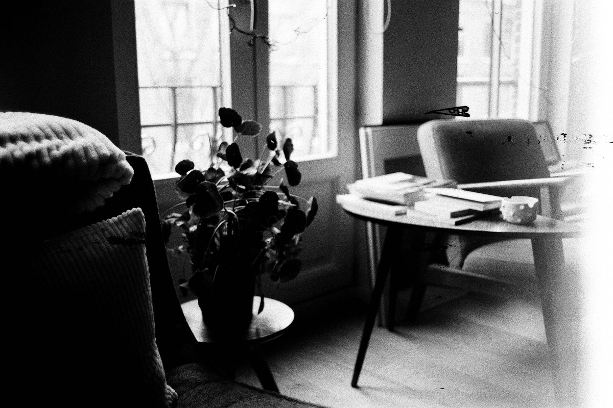 The image displays a cozy living room. A couch and coffee table are visible, with items like books and a plant on the table. A window with blinds lets