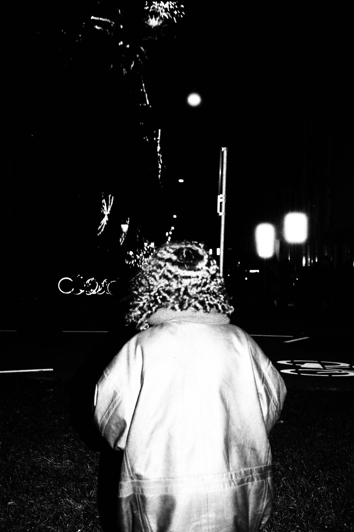 This is a black and white photo of a child standing on the side of a street. The child appears to be wearing a raincoat and looking away from the came