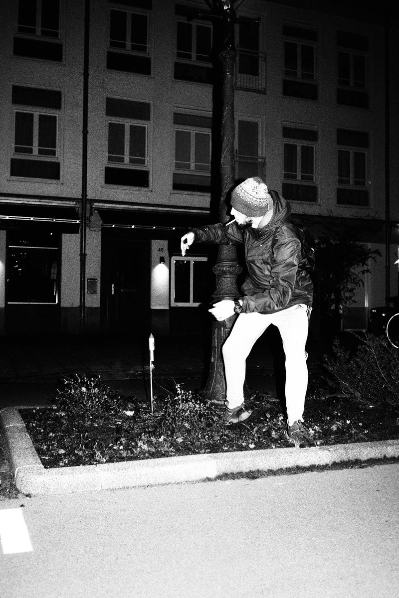 Black and white image of man standing next to a small tree. He is wearing a beanie, jacket, and jeans. The man is looking down at the ground. Behind h