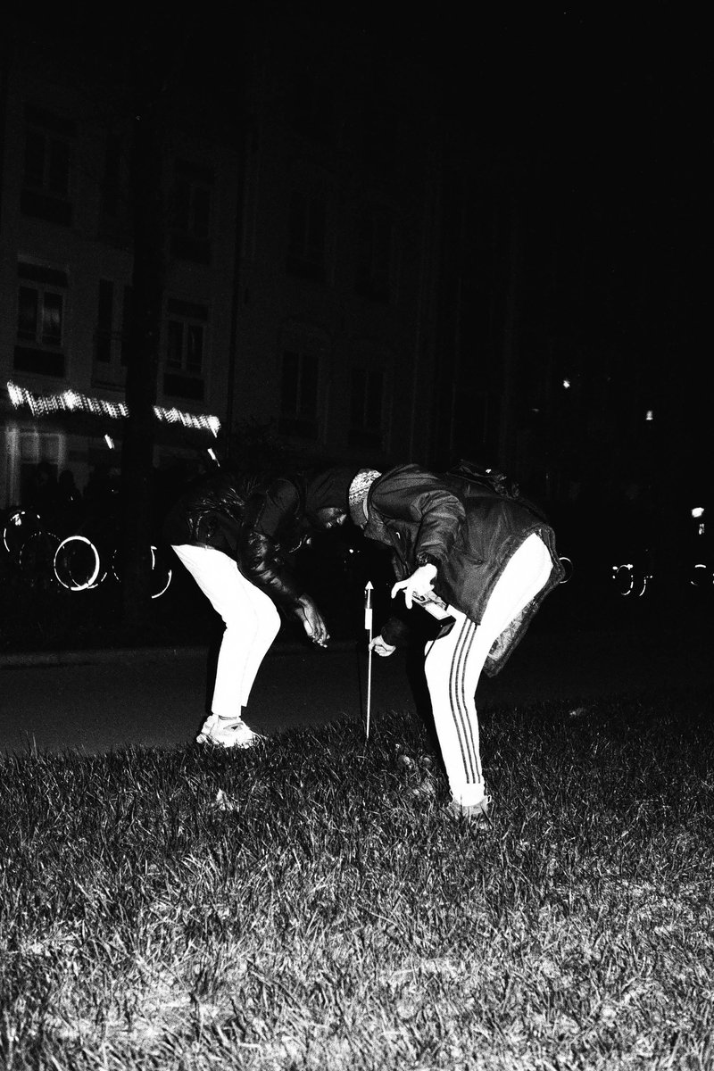 Two people playing golf at night on the street. The man is crouched while the woman stands in front of him, both with clubs. They are dressed casually