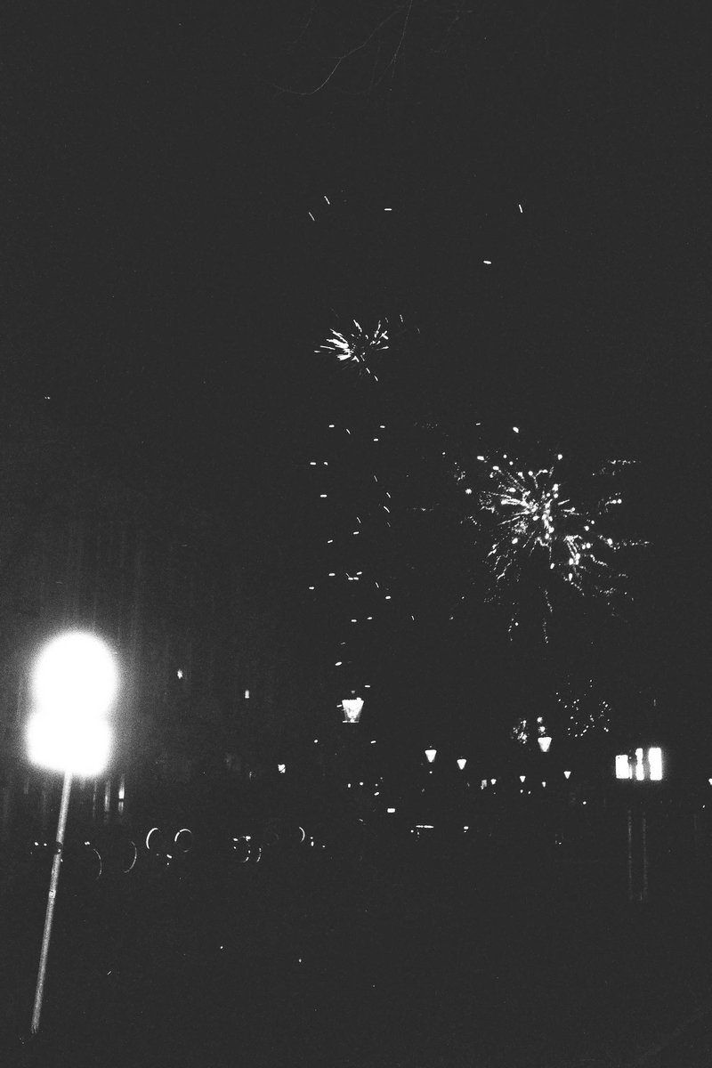 The image is a black and white photo capturing a lively night scene. At the center of the frame, there's a fireworks display with explosions lighting