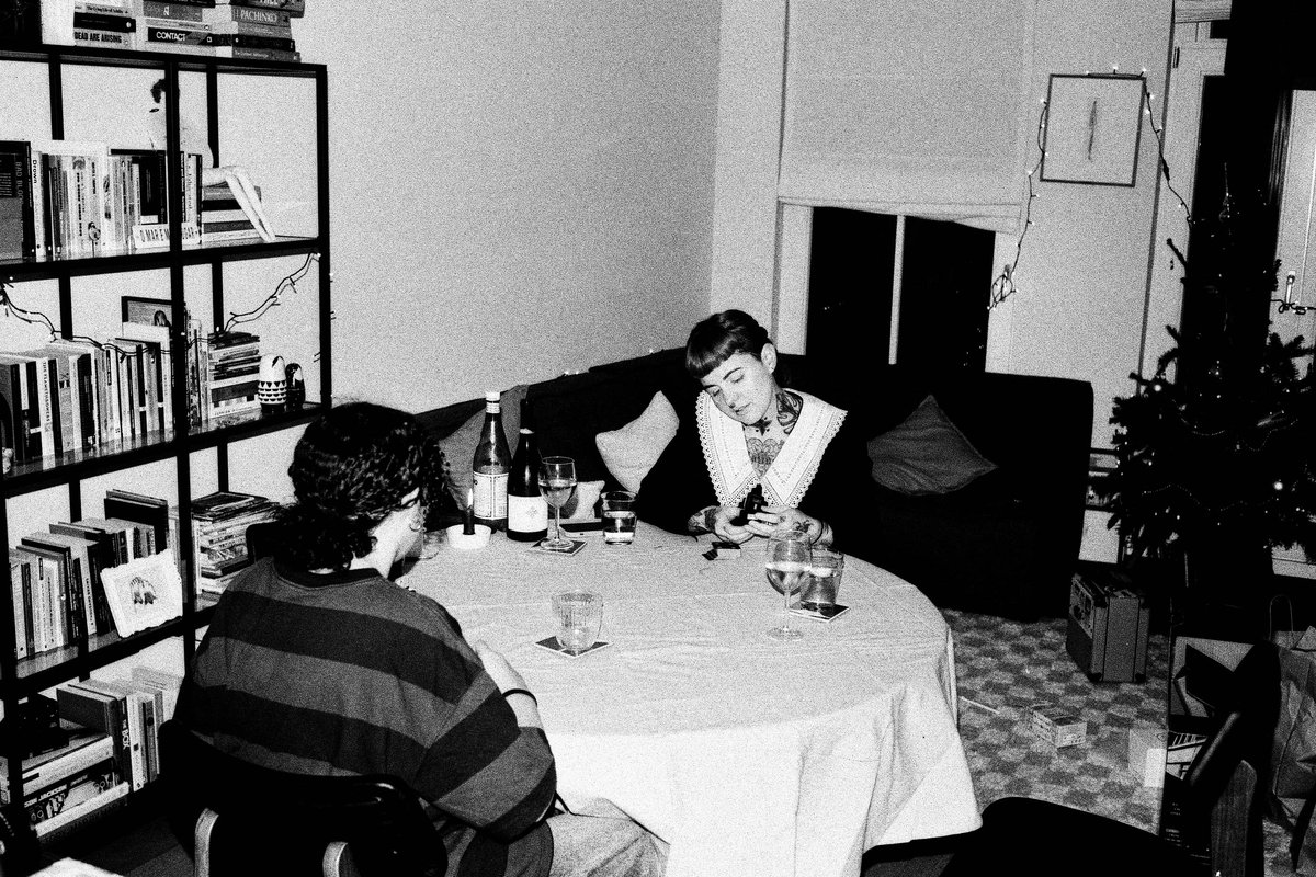 The image is a black and white photograph featuring a man standing at a counter. He is holding a bottle of wine and appears to be inspecting or smelli
