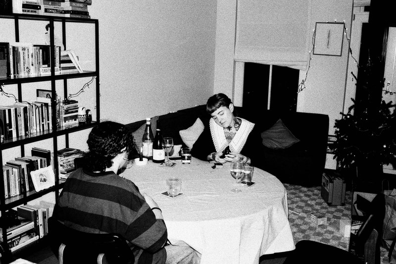 The image is a black and white photograph featuring a man standing at a counter. He is holding a bottle of wine and appears to be inspecting or smelli