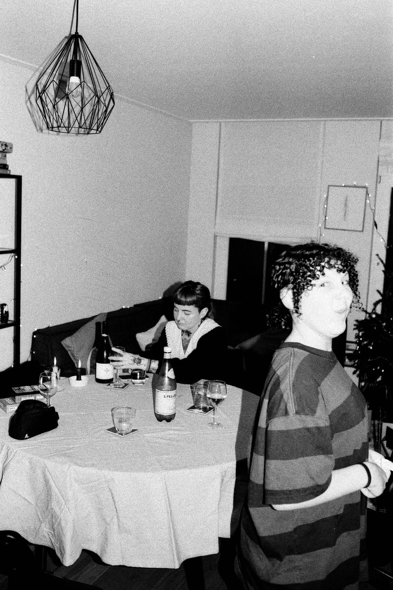 The image is a low-resolution black and white photo of a man standing in the center of a room. He has dark hair and appears to be looking directly at