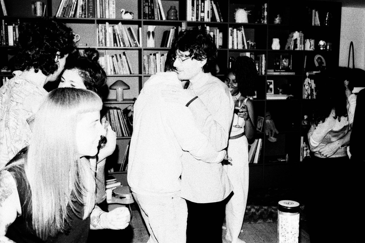 The image is a black and white photo of three men posing. In the background, there's a partial view of furniture and what appears to be a television s