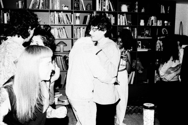 The image is a black and white photo of three men posing. In the background, there's a partial view of furniture and what appears to be a television s