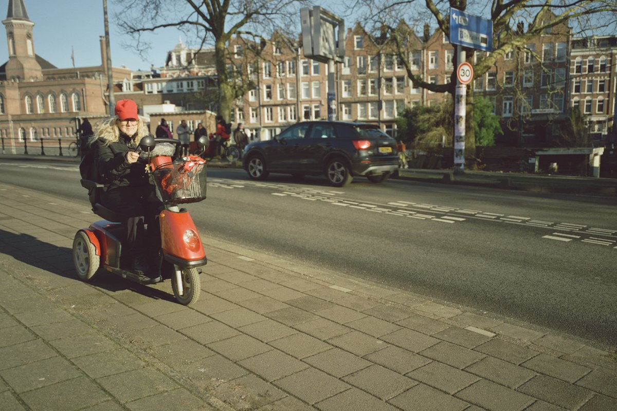  The image shows an urban street scene with a person riding a motor scooter on the road. The rider appears to be wearing a red helmet and is moving down the street. There are buildings visible, indicating a residential or commercial area. Additionally, there are vehicles and pedestrians present in the scene, suggesting it's a busy time of day. The photo seems to have been taken during the day under natural lighting conditions. 