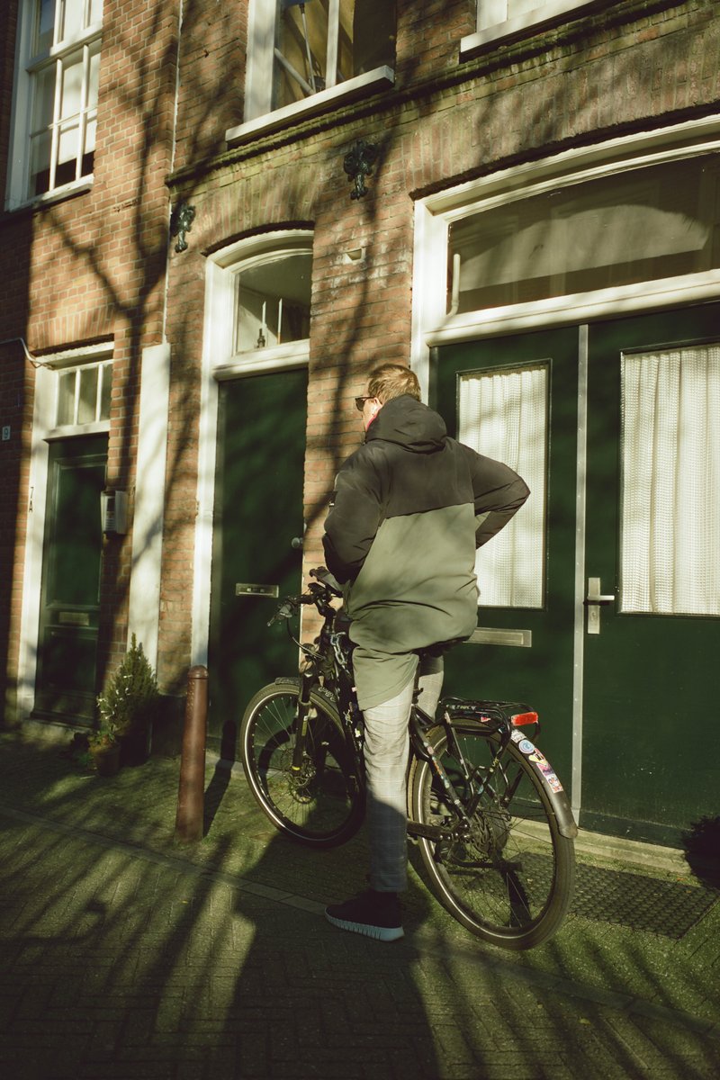 The image shows a young man standing outside a building with a bicycle parked next to him. He is wearing casual clothing, and the setting appears to be an urban environment with brick buildings and trees lining the street.