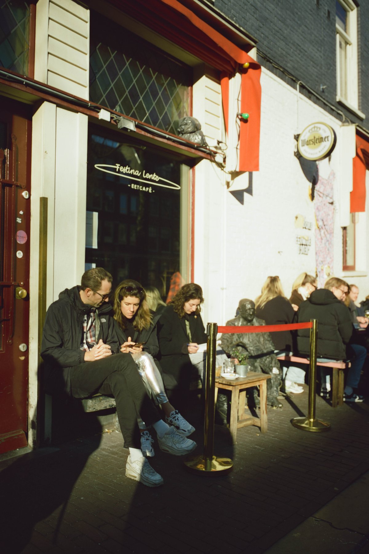  The image depicts an outdoor café scene with people sitting at tables outside. There are individuals seated under shaded areas, engaged in various activities such as using cell phones or dining. The setting suggests a casual urban environment with a relaxed atmosphere, indicated by the presence of benches and potted plants. Businesses with colorful signs are visible on the street-side background, contributing to the bustling city vibe. 