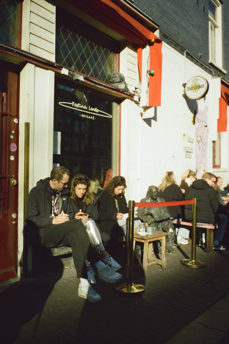 The image depicts an outdoor café scene with people sitting at tables outside. There are individuals seated under shaded areas, engaged in various activities such as using cell phones or dining. The setting suggests a casual urban environment with a relaxed atmosphere, indicated by the presence of benches and potted plants. Businesses with colorful signs are visible on the street-side background, contributing to the bustling city vibe.