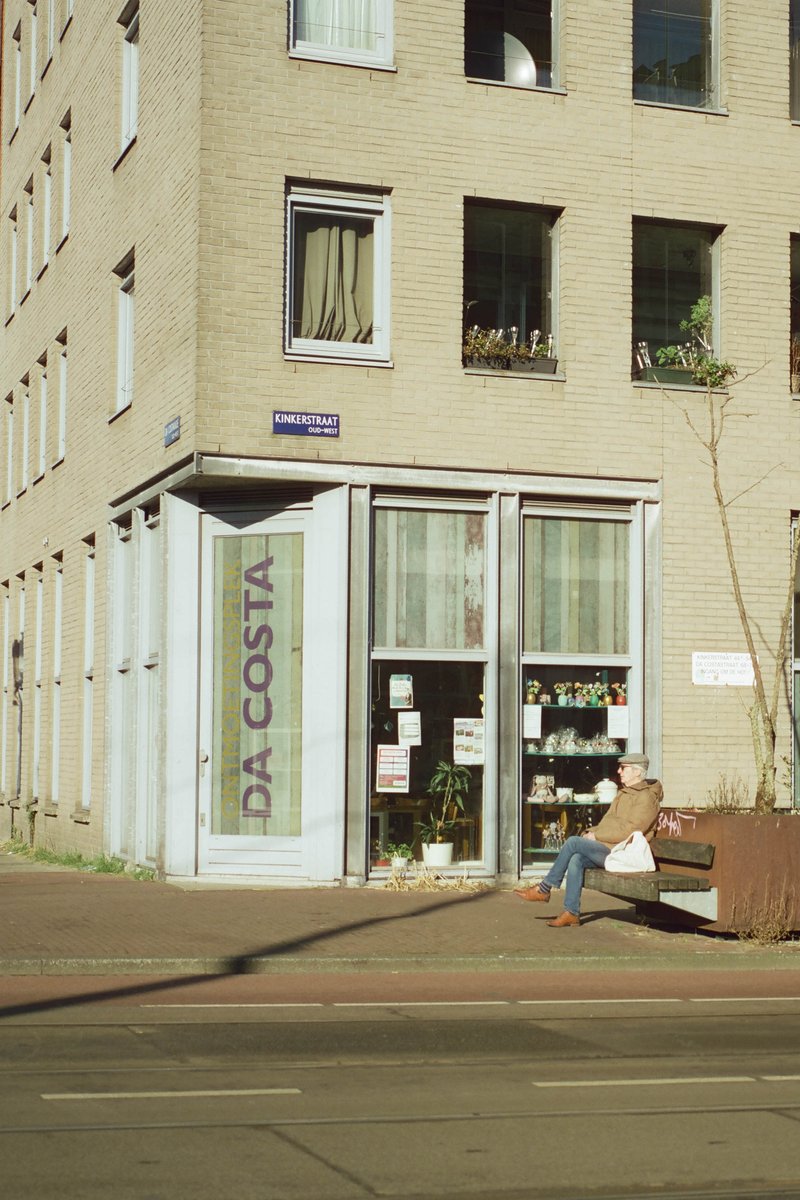 The image shows an urban street scene with a two-story building prominently featuring a storefront named "Da Costas". Outside of the store, there is a person seated on a bench. On the first floor of the building, a couple of windows are visible with curtains partially drawn. The ground floor window displays various items, possibly merchandise for sale. The sky appears overcast, and the street has a few other individuals scattered along its length.