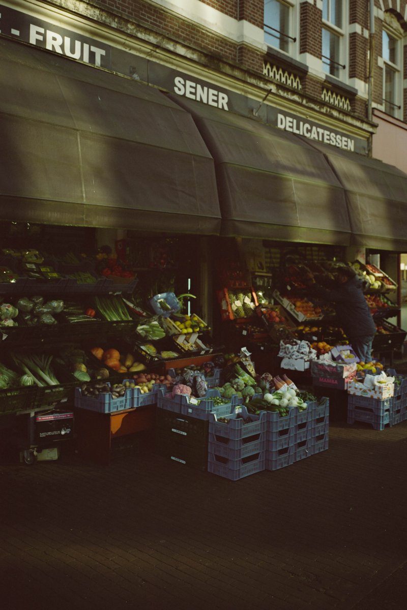 The image captures a vibrant outdoor farmers market bustling with fresh produce. Various fruits and vegetables are neatly arranged in crates, their colors popping against the backdrop of the market stall. The stall itself is covered by a large umbrella, providing shade for the produce and possibly customers or vendors. There's a sign in the background that reads "Fruit & Vegetable Deli", indicating the nature of the establishment. The overall scene suggests a lively community gathering place where people can purchase fresh, locally grown items.