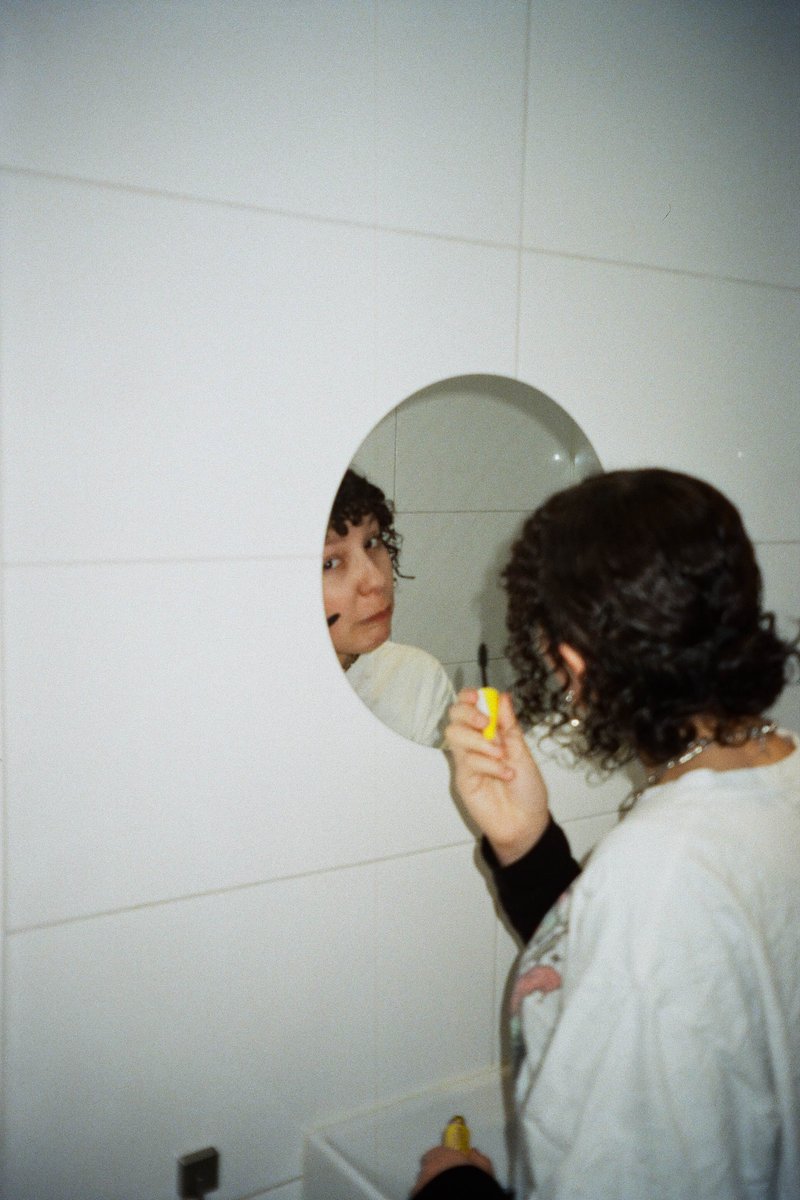 The image shows an indoor scene where two individuals are present. One person is standing close to a wall-mounted mirror, appearing as if they are being reflected in it. This person is holding a yellow object and has short hair. There's another individual nearby who seems to be looking at the first person through a small circular window or portal. The background features plain walls and what appears to be a countertop or ledge with a few items on it, including a bottle.