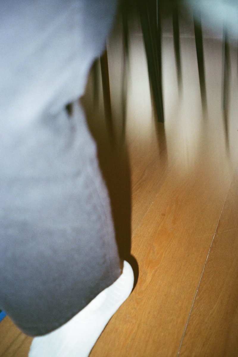 The image shows a person standing in front of a wooden floor, with a dimly lit area visible in the background. The individual is wearing dark clothing and their feet are visible but only one sock can be seen. The lighting in the room creates an atmospheric effect, suggesting it might be nighttime or in a low-light environment.