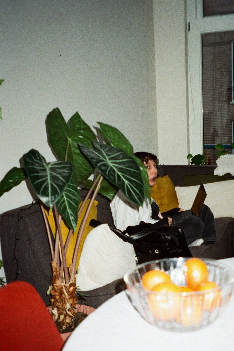 In the image, there is a cozy indoor setting with a person sitting on a couch and looking at a potted plant nearby. The person is dressed in a yellow top. The room has a homely vibe, with a window covered by blinds in the background. On the table next to the couch, there are a couple of books and an orange, suggesting that someone might have been reading or eating while relaxing on the couch.