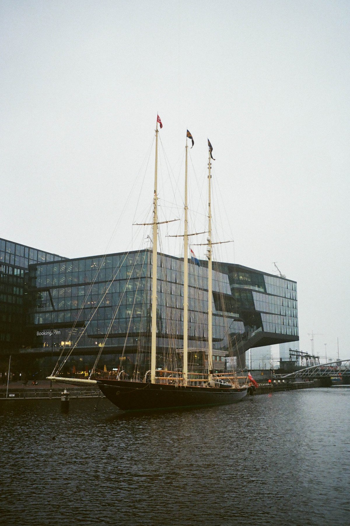  The image depicts a large sailing ship docked at a pier. The ship is the central focus of the scene, with its masts, rigging, and sails visible against a backdrop of buildings. There are no people immediately around the ship, but there's an air of activity on the dock, suggesting a bustling harbor environment. The image has a sepia tone that gives it a timeless quality. 