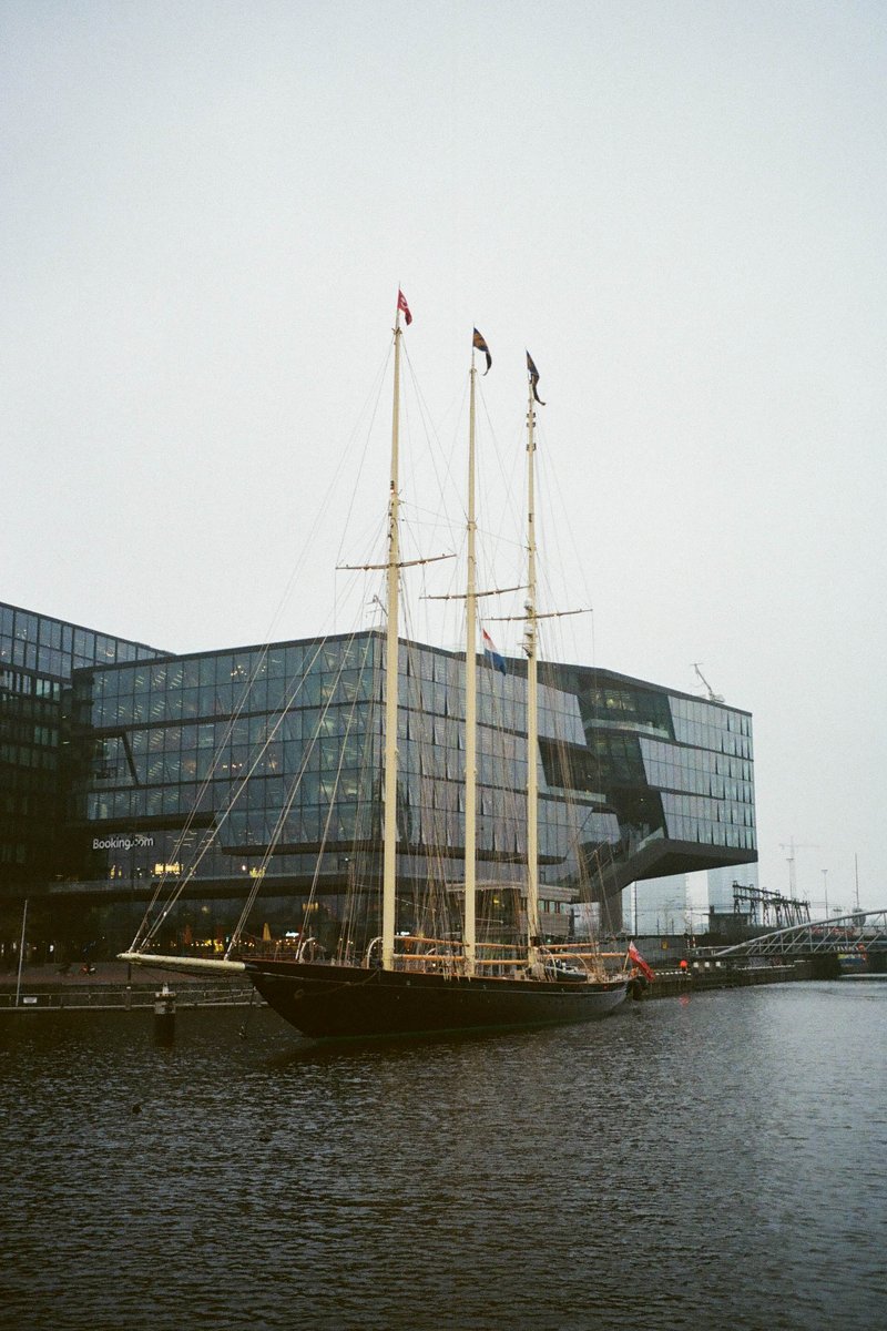 The image depicts a large sailing ship docked at a pier. The ship is the central focus of the scene, with its masts, rigging, and sails visible against a backdrop of buildings. There are no people immediately around the ship, but there's an air of activity on the dock, suggesting a bustling harbor environment. The image has a sepia tone that gives it a timeless quality.