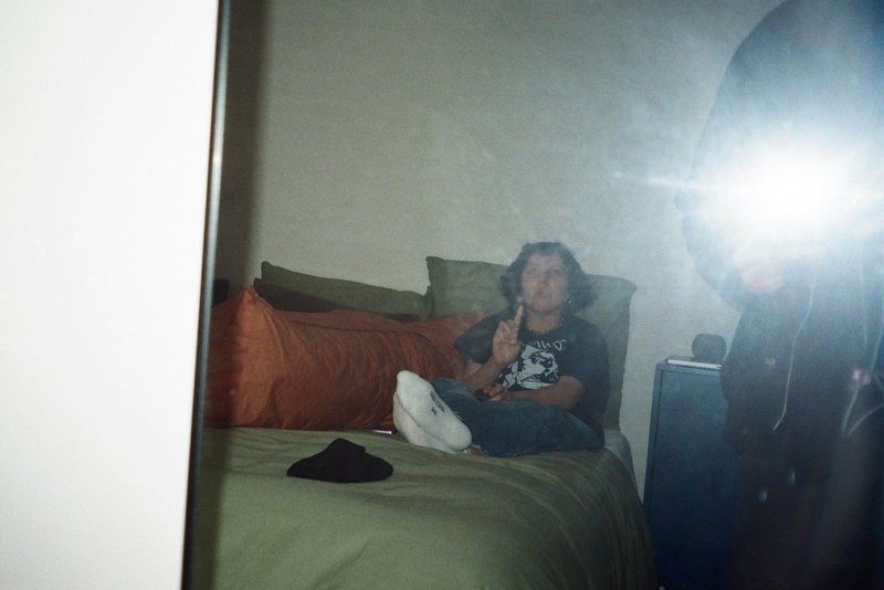 The image features an individual sitting on a bed with a cell phone in front of them. They appear relaxed, casually dressed, with one hand resting on the bed and the other holding the phone. In the foreground, there's a mirror reflecting the person's surroundings and another image captured through it. The room has a simple decor, with an orange pillow providing a splash of color against the neutral tones of the walls. The overall setting suggests a quiet moment at home or in a hotel room.