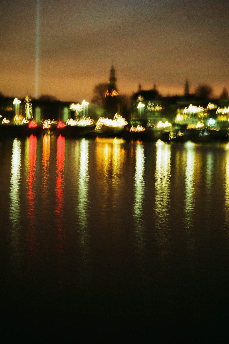 The image captures a nighttime scene of a cityscape, with a large body of water reflecting the lights of buildings across the way. The water is calm, and on it, boats are visible. The sky above the water is dark, suggesting late evening or night time. A town can be seen in the distance.