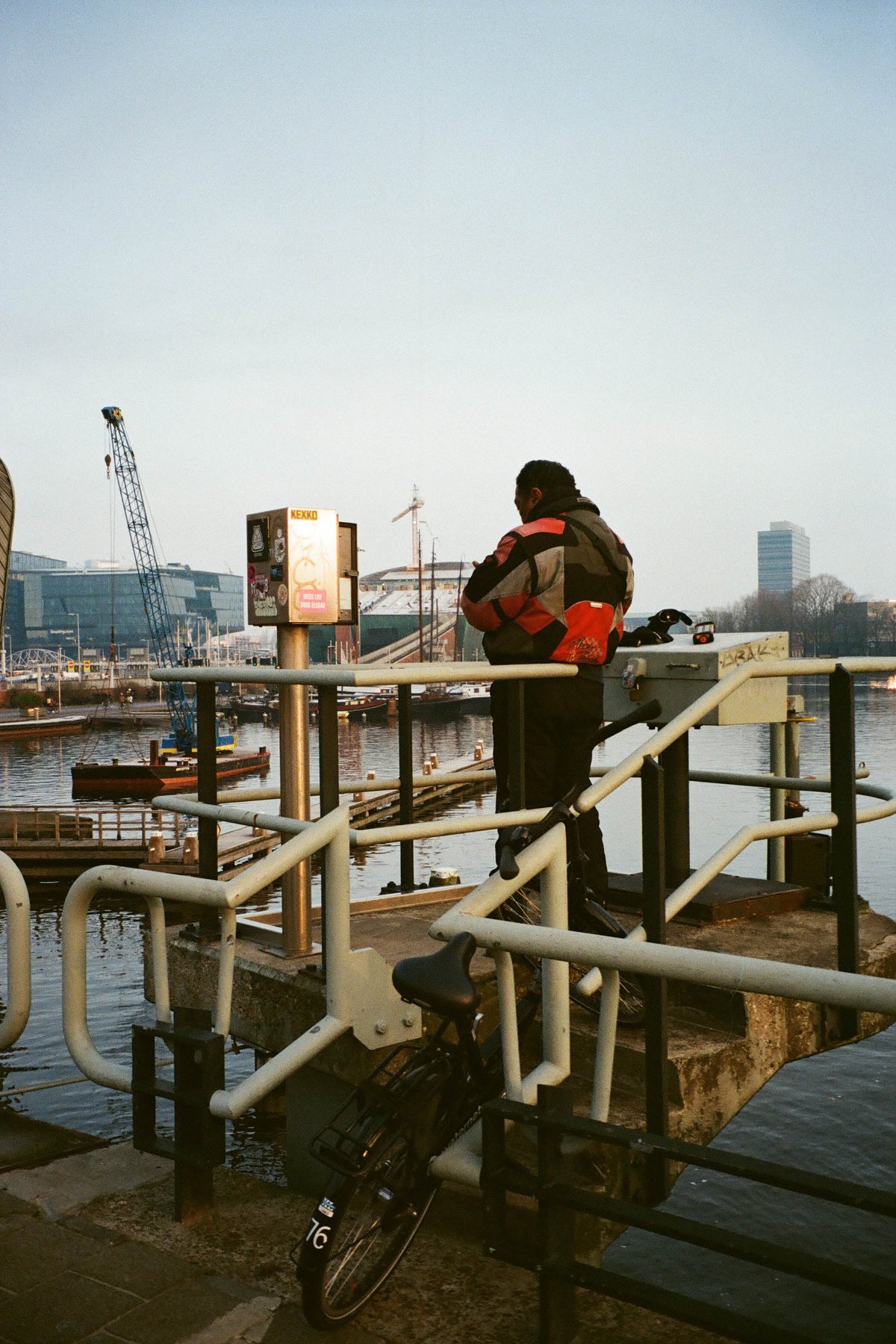  The image depicts a person standing on a dock by a body of water, holding an item and seemingly looking at something out of frame. In the background, there's a bridge with a crane, indicating a construction or industrial area nearby. The sky is overcast, suggesting it might be either early morning or late afternoon. 