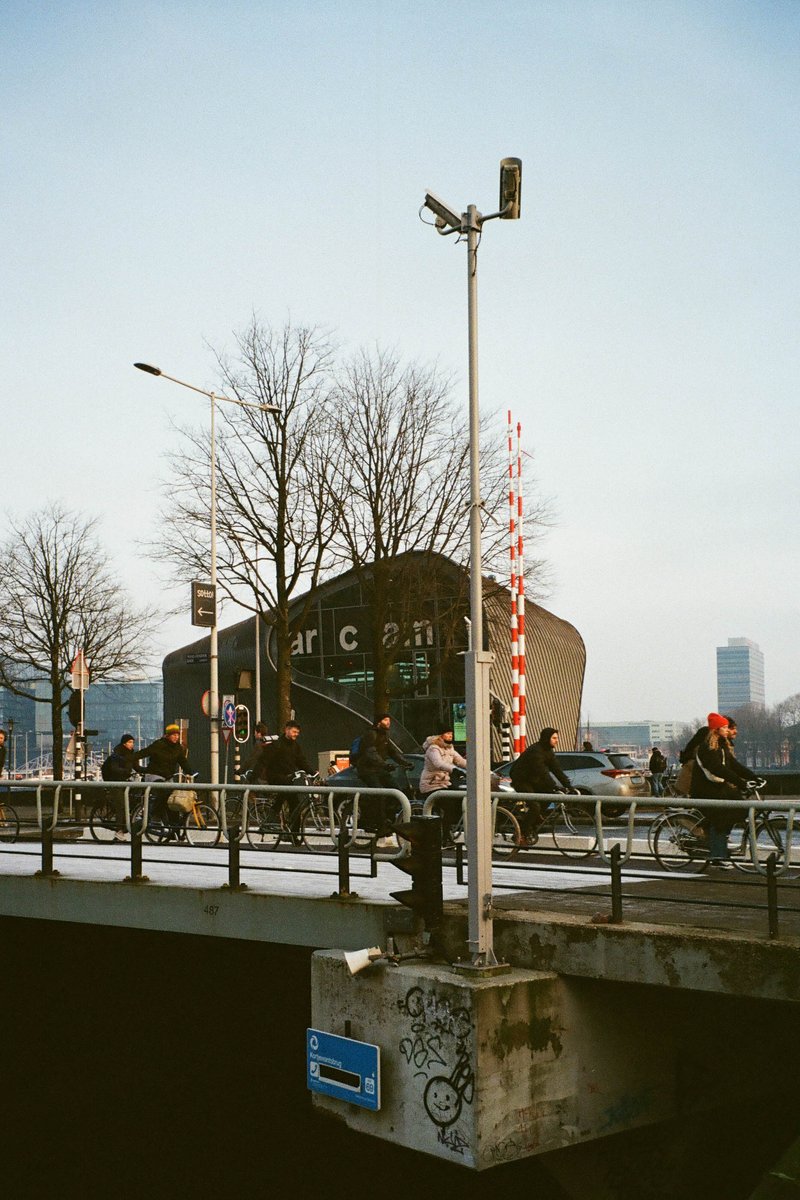 The image shows an outdoor scene with a bridge over a body of water. A street light is prominently featured in the center, casting shadows and highlighting its presence. There are people riding bicycles on the bridge, indicating movement and activity. In the background, there's a building with a distinctive architectural feature, possibly a church or another type of public building. The sky is clear, suggesting it might be daytime.